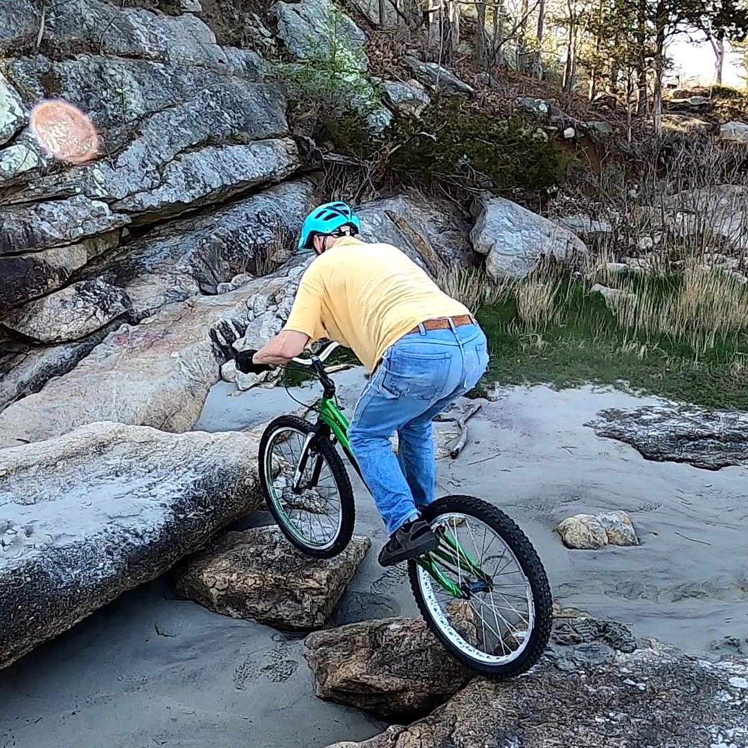 Jim rides his trials bike across a line of rocks to a boulder, trying to avoid the sand below