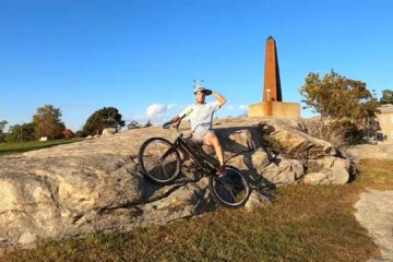 Tony sits on a rock with his bike, saluting us