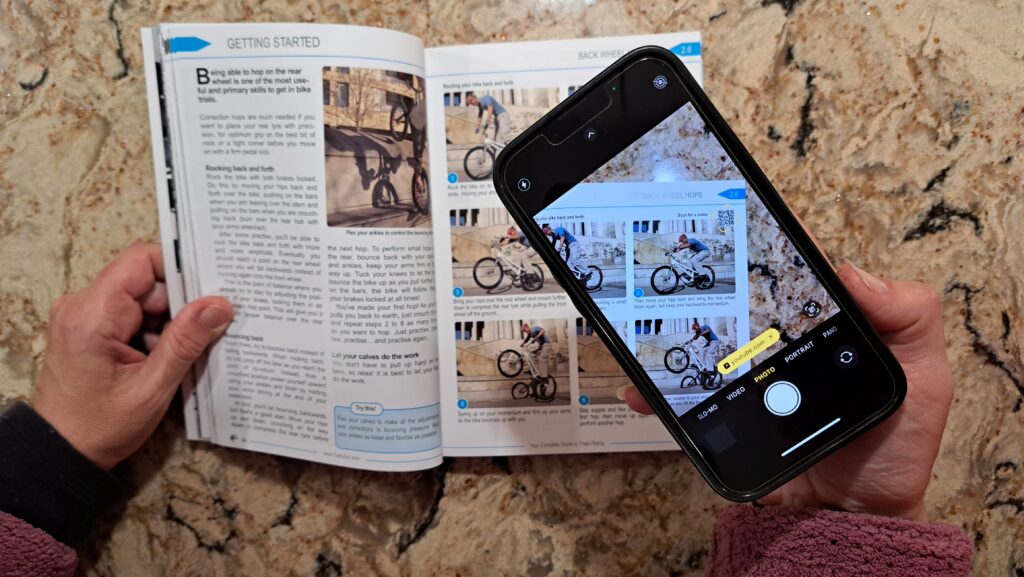 Smartphone being used to access a YouTube video (back wheel hops) using a QR code embedded in the book Your Complete Guide To Trials Riding - Street Trials Augmented Edition, by Julien Happich